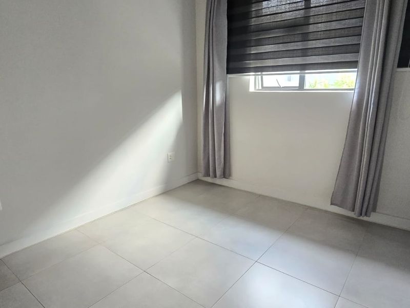 A bright, empty bedroom featuring neutral tiled flooring, clean white walls, and a window with fitted blinds.