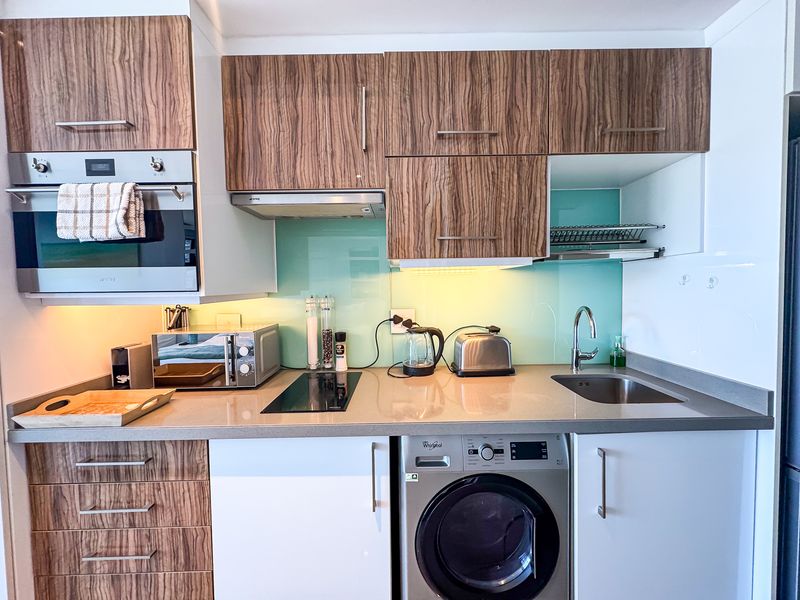 Modern compact kitchen featuring wood-grain and white cabinetry, stone countertops, glass splashback, built-in oven, and an integrated washing machine.