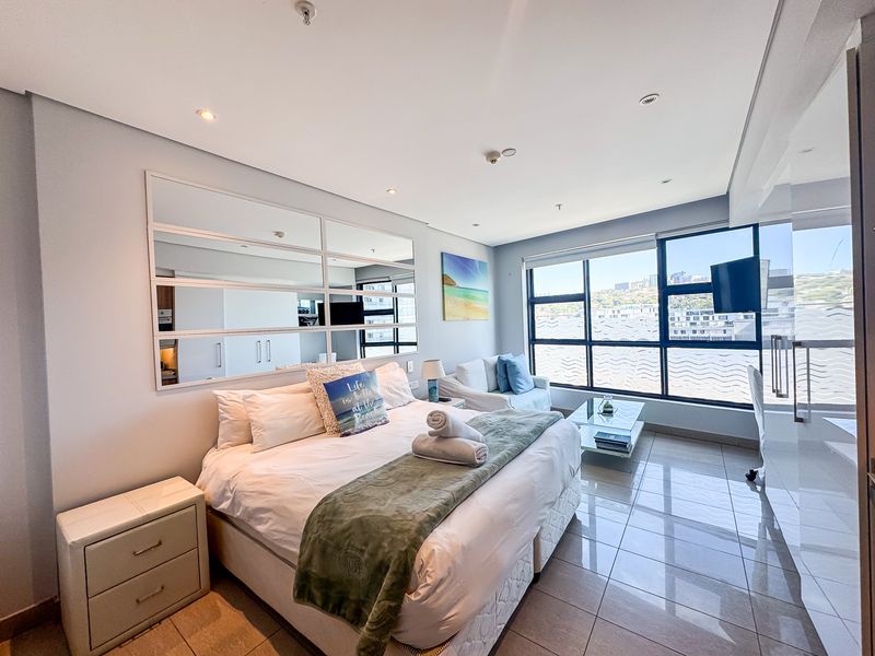 A bright and modern bedroom featuring large format floor tiles, recessed lighting, and expansive windows offering natural light and urban views.