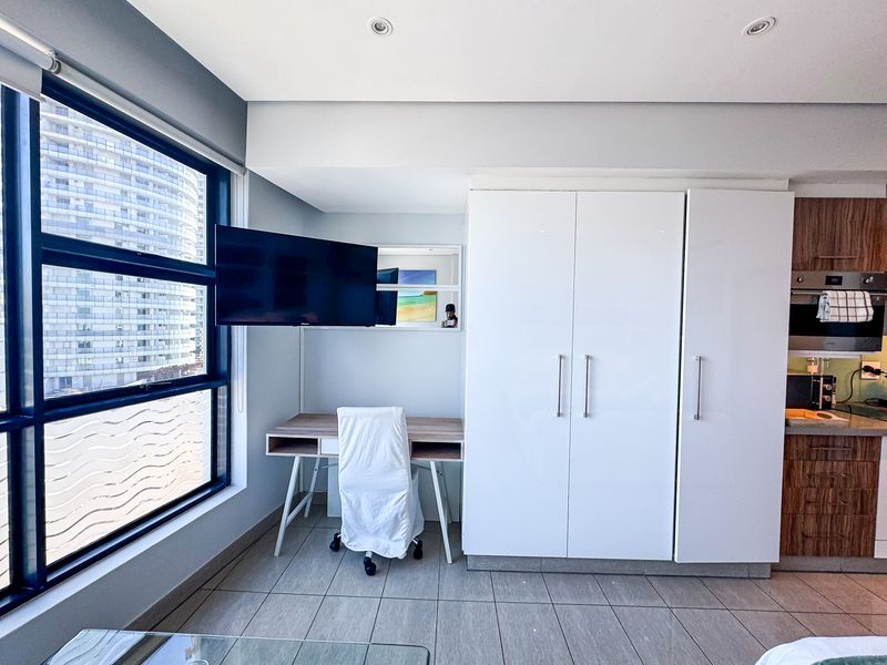 Modern open-plan apartment area featuring built-in cabinetry, integrated kitchen appliances, tiled flooring, and large windows providing ample natural light.