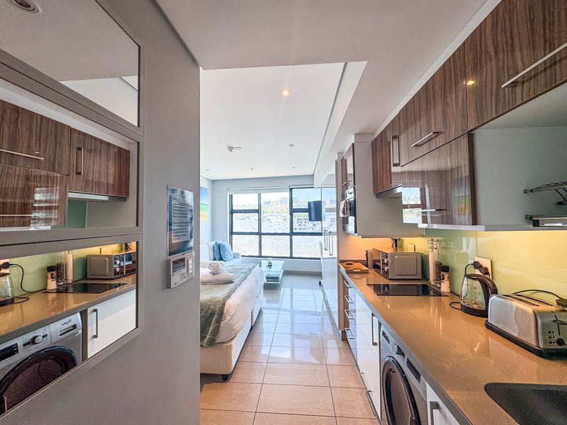 Modern open-plan apartment featuring a sleek kitchen with integrated appliances and stone countertops, flowing seamlessly into a bright bedroom area with large windows.