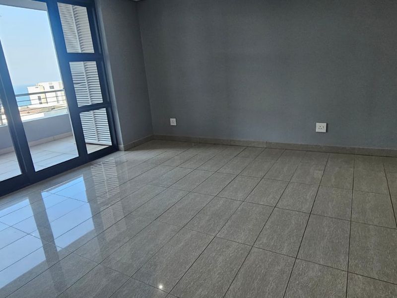 A bright, empty living area featuring polished tile flooring and large sliding glass doors that open onto a balcony with coastal views.