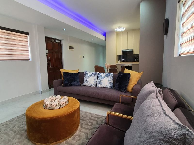 Modern open-plan living area featuring recessed ceiling lighting, LED accent strips, tiled floors, and a compact, well-equipped kitchen with built-in cabinetry and appliances.