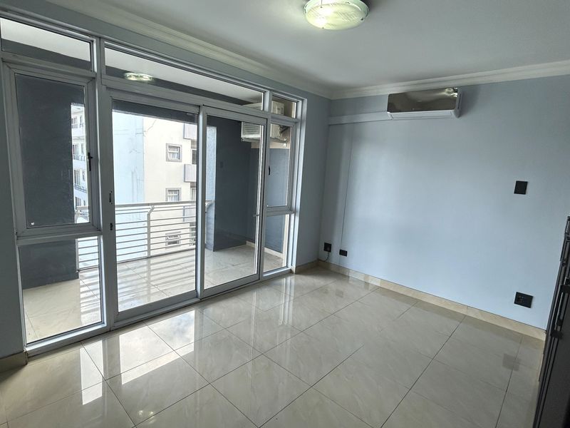A bright, empty living room featuring large sliding glass doors leading to a balcony, tiled flooring, and a wall-mounted air conditioning unit.