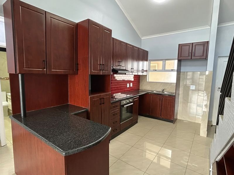 A well-maintained kitchen featuring dark wood cabinetry, granite countertops, built-in oven and hob, and tiled flooring, adjacent to an internal staircase.