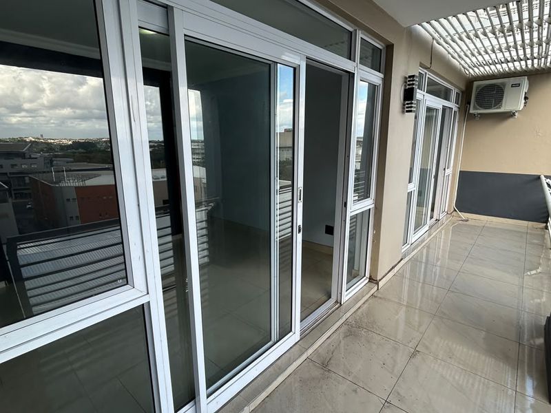 A spacious covered balcony featuring tiled flooring, a slatted pergola roof, and sliding glass door access to the interior living area.