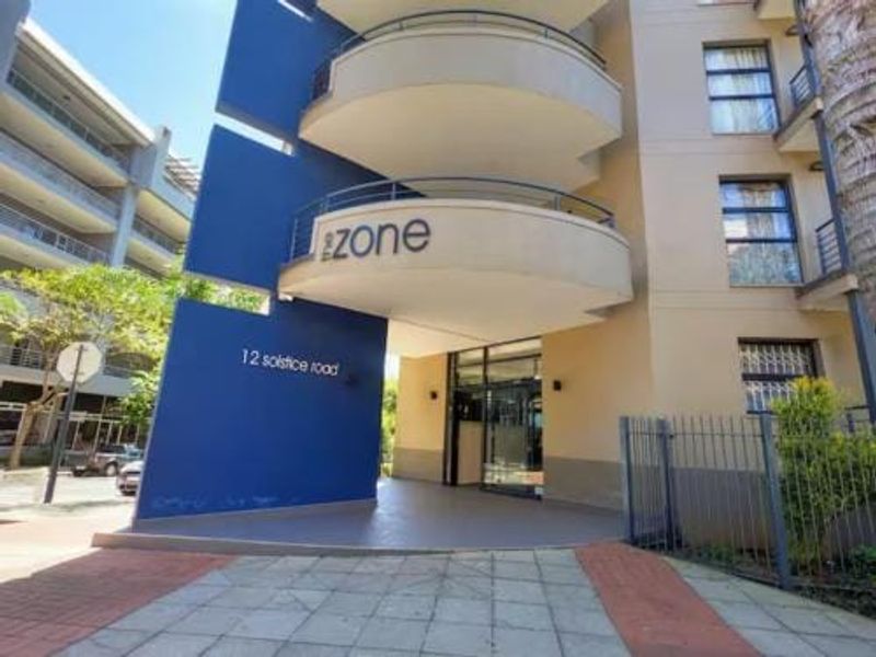 Front entrance view of The Zone apartment complex, featuring modern blue architectural accents, curved balconies, and a paved pedestrian access area.