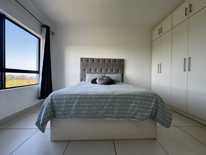 A bright bedroom featuring neutral tiled flooring, ample built-in cupboard storage, and a large window offering natural light and scenic views.