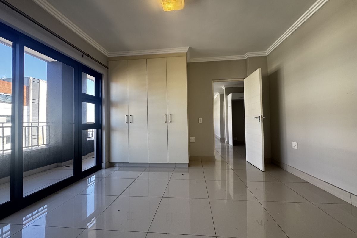 A bright, empty bedroom featuring neutral tiled flooring, built-in cupboards, and a sliding glass door providing access to a private balcony.