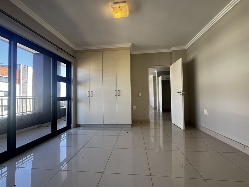 A bright, empty bedroom featuring neutral tiled flooring, built-in cupboards, and a sliding glass door providing access to a private balcony.