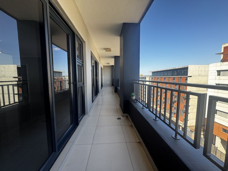 A spacious, modern apartment balcony featuring tiled flooring, a secure metal balustrade, and large sliding glass doors providing access to the interior.
