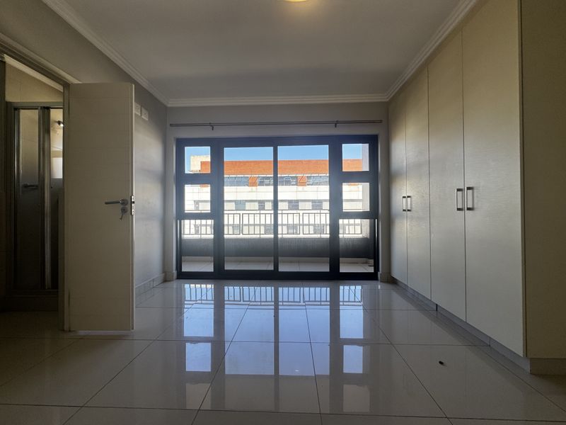 A bright, empty bedroom featuring tiled floors, extensive built-in cupboards, and a large sliding glass door providing access to a private balcony.