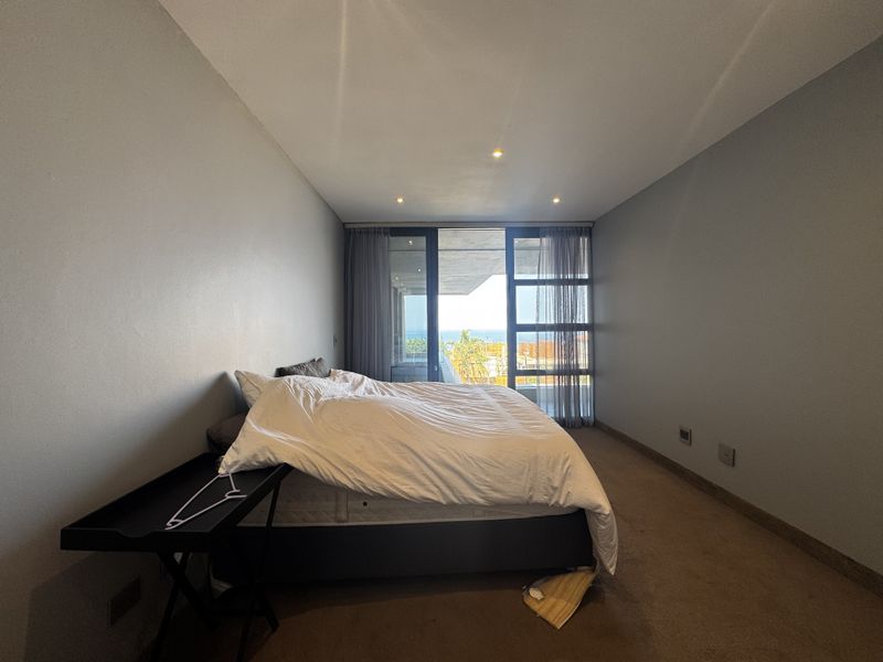 A spacious bedroom featuring neutral walls, carpeted flooring, recessed lighting, and large sliding glass doors providing access to a balcony with ocean views.