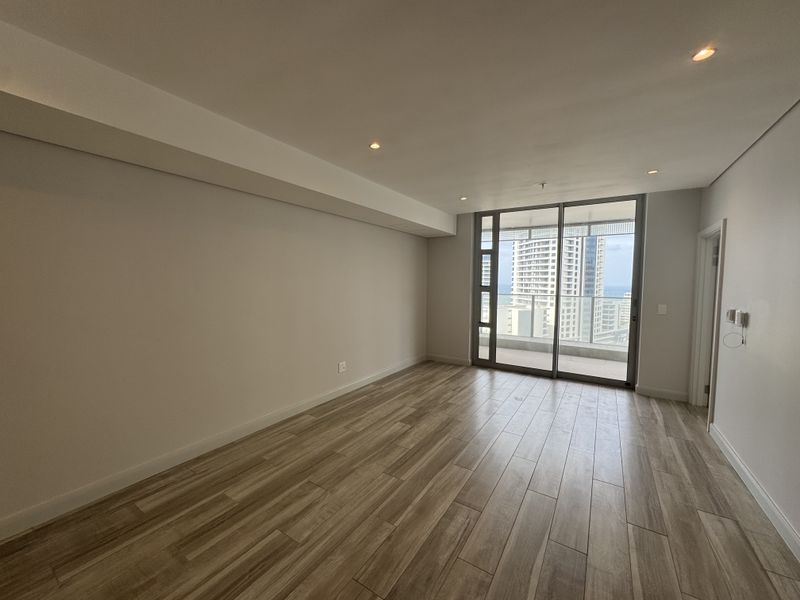 A bright, empty living area featuring modern wood-look tile flooring, recessed lighting, and large sliding glass doors leading to a balcony with city views.