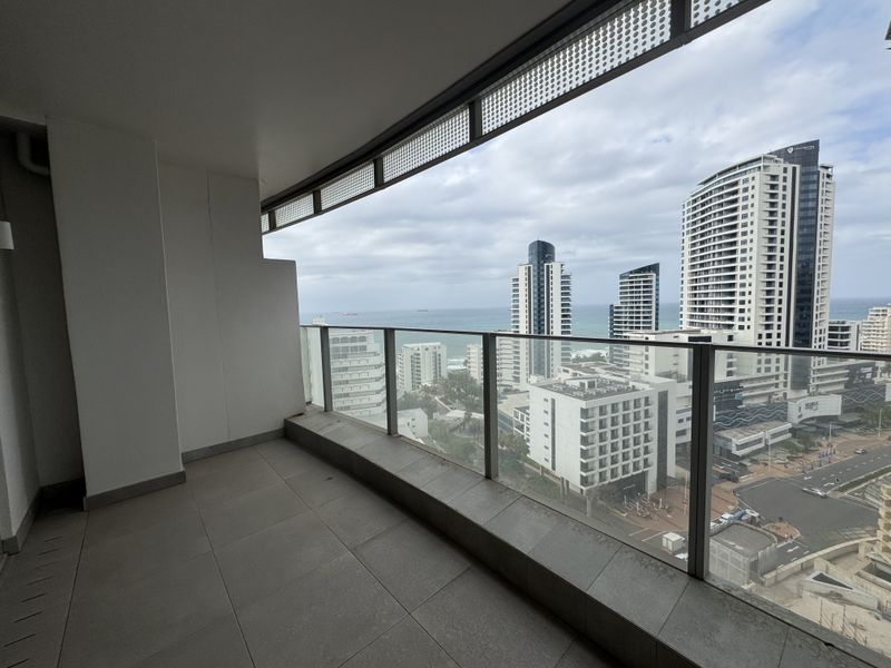 A spacious covered balcony featuring a modern glass balustrade and tiled flooring, offering expansive urban and coastal views.