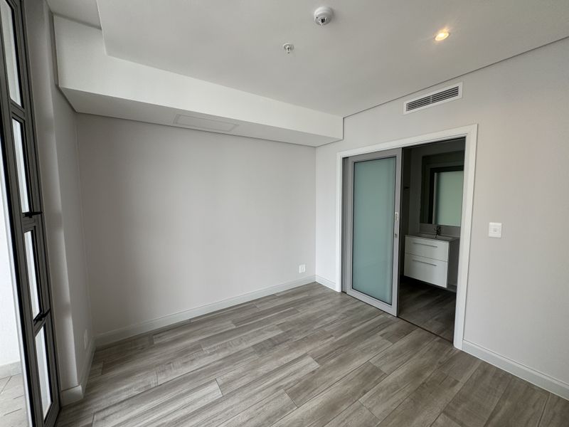 A bright, modern bedroom featuring wood-look laminate flooring, recessed lighting, and a sleek sliding door leading to an en-suite bathroom.