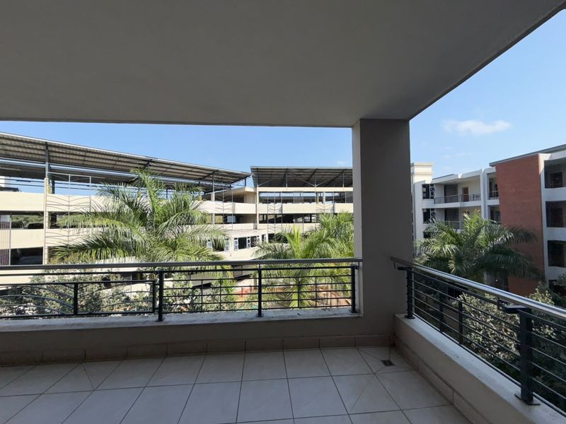A spacious covered balcony featuring tiled flooring and a metal balustrade, offering an open view of the surrounding apartment complex and greenery.