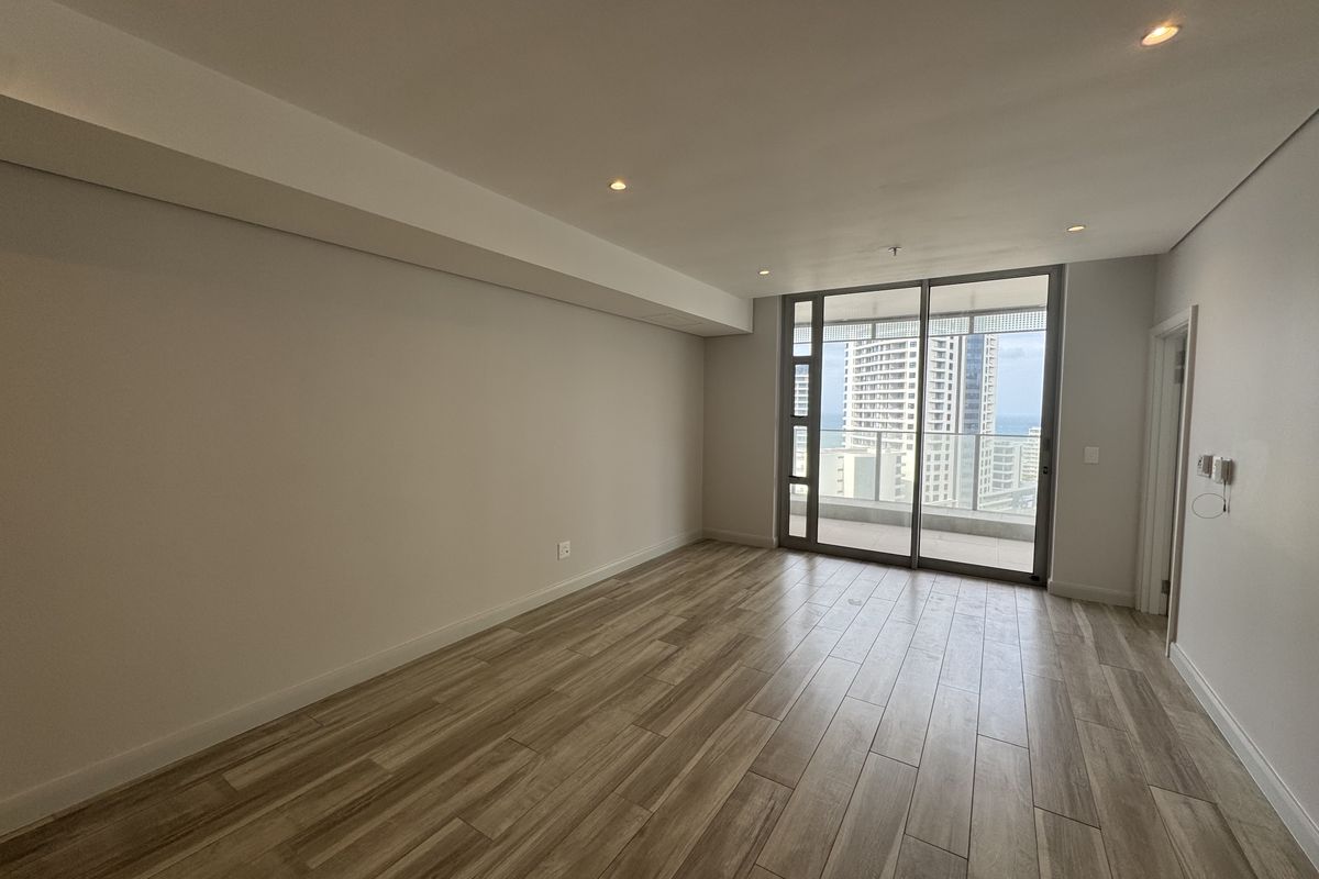 A bright, empty living area featuring modern wood-look tile flooring, recessed lighting, and large sliding glass doors leading to a balcony with city views.