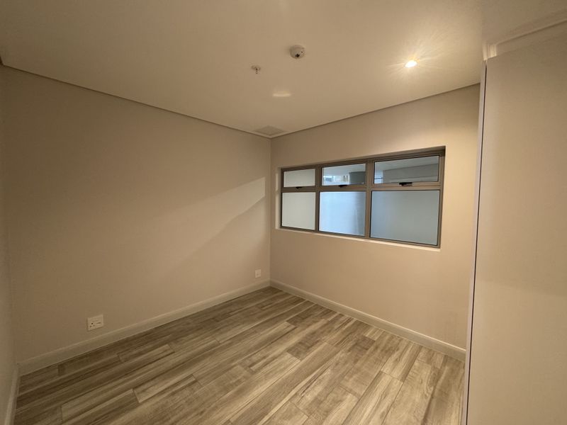 A clean, empty bedroom featuring modern laminate flooring, neutral walls, and an aluminium-framed window, providing a bright and versatile space.
