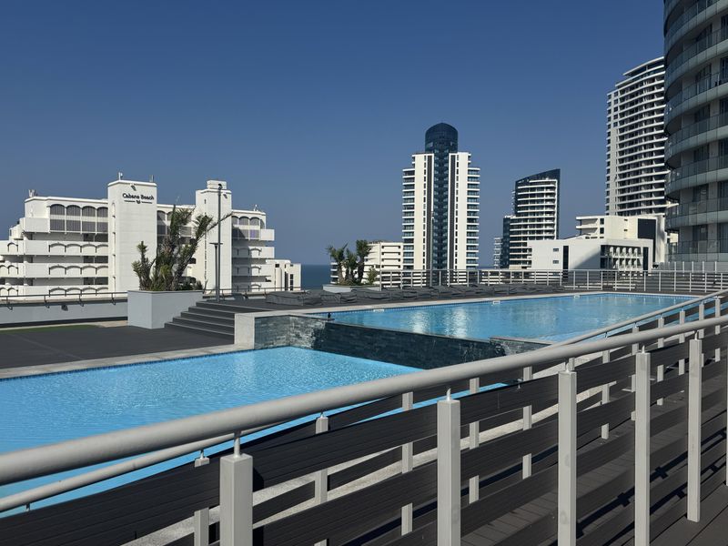 A modern elevated swimming pool area featuring tiered decking, stone water features, and metal safety railings with surrounding urban views.
