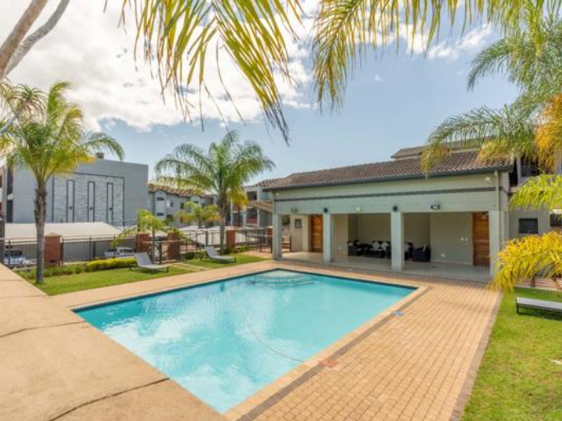 Communal pool area featuring a swimming pool, paved deck, and a covered patio space within a landscaped garden setting.