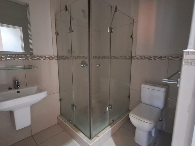 A clean, functional bathroom featuring a pedestal washbasin, a glass-enclosed corner shower, and a standard toilet with neutral wall and floor tiling.