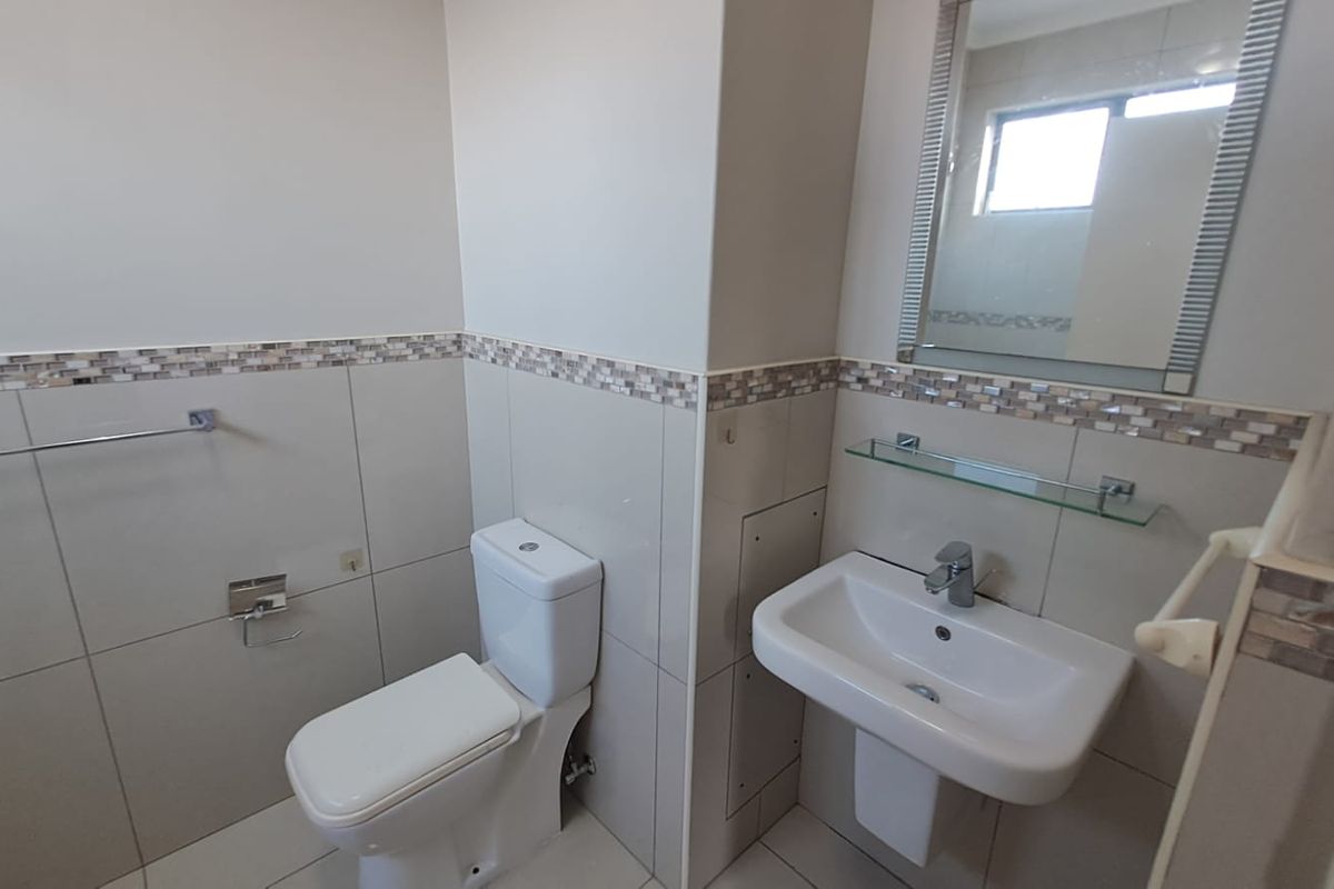 A clean and functional bathroom featuring neutral ceramic wall tiles with a decorative mosaic border, a pedestal basin, and a wall-mounted mirror.