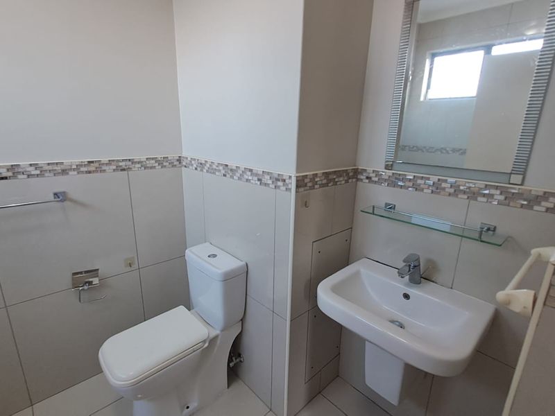 A clean and functional bathroom featuring neutral ceramic wall tiles with a decorative mosaic border, a pedestal basin, and a wall-mounted mirror.