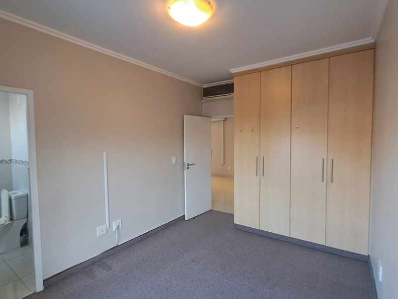 A spacious bedroom featuring neutral walls, carpeted flooring, and ample built-in cupboard storage, with an adjacent en-suite bathroom visible.