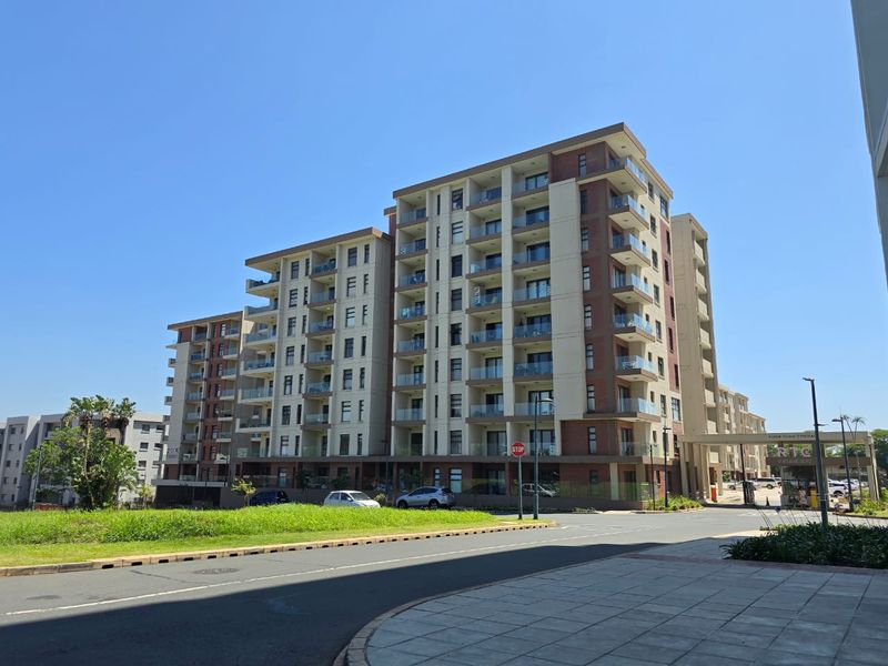 A modern multi-story apartment complex featuring brick and concrete facade, glass-fronted balconies, and a paved entrance area with a security gatehouse.