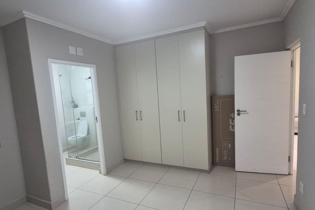 A clean, neutral bedroom featuring tiled flooring, built-in cupboards, and an en-suite bathroom with a glass shower enclosure.