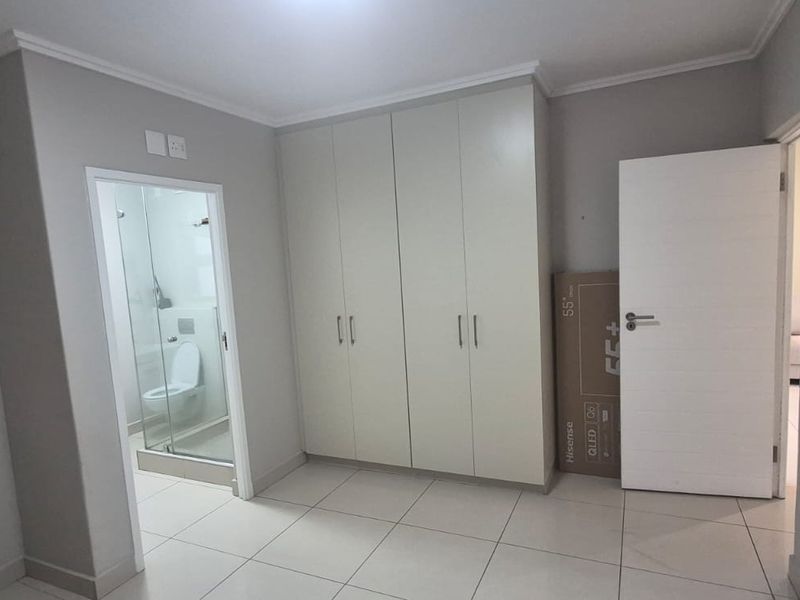 A clean, neutral bedroom featuring tiled flooring, built-in cupboards, and an en-suite bathroom with a glass shower enclosure.