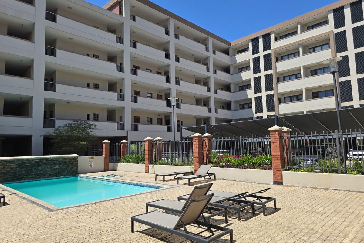 Communal pool area featuring a paved deck, water feature, and shade structure, set within a modern apartment complex courtyard.