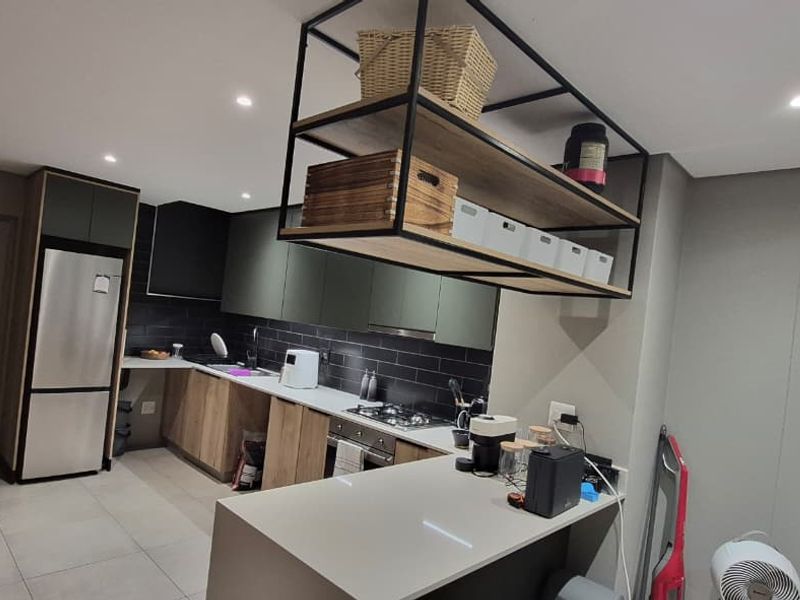 Modern kitchen featuring dark cabinetry, stone countertops, a gas hob, and a unique suspended metal shelving unit, complemented by an exposed brick feature wall.