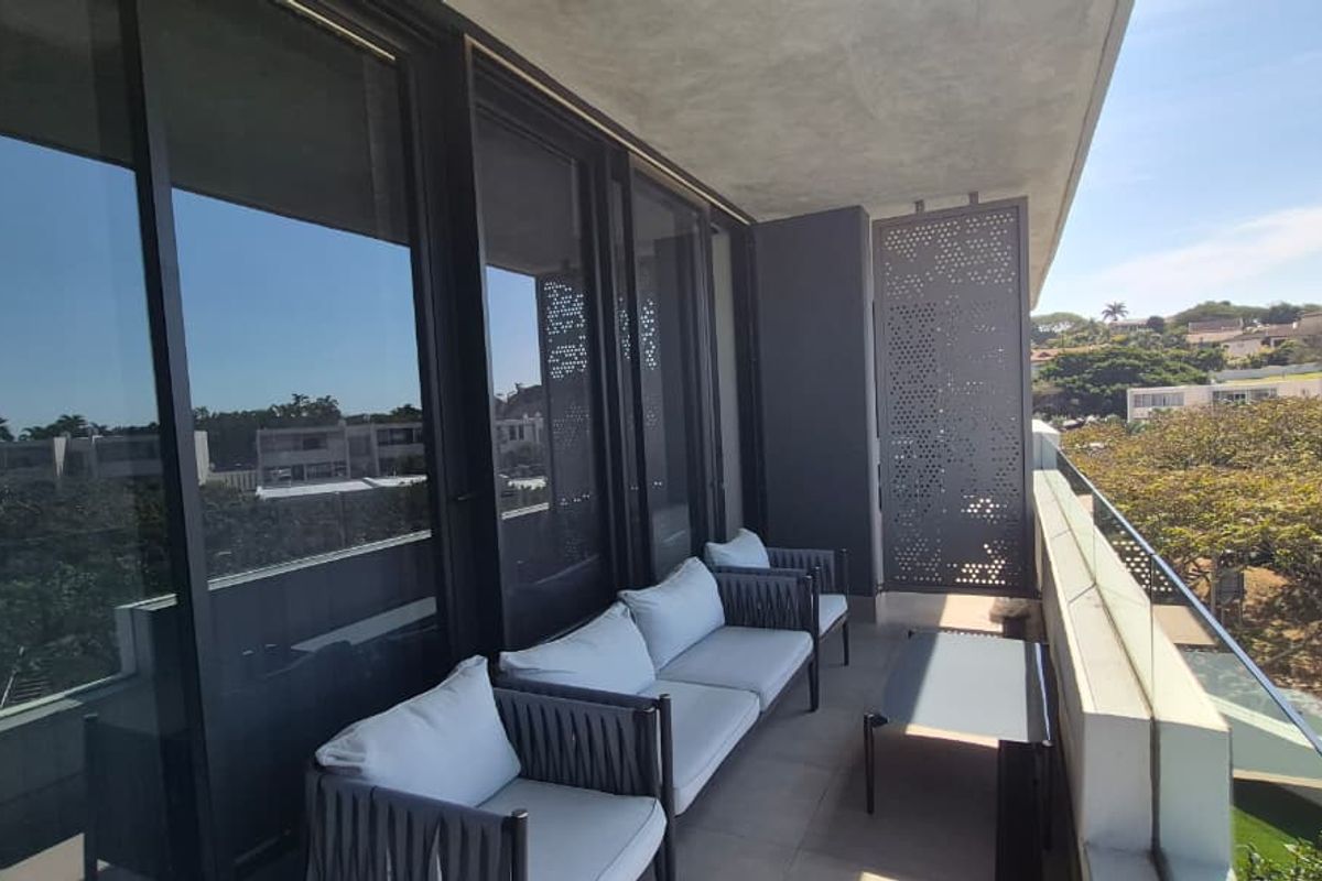 A modern covered balcony featuring a glass balustrade, sleek sliding doors, and a decorative perforated metal privacy screen overlooking the surrounding neighborhood.