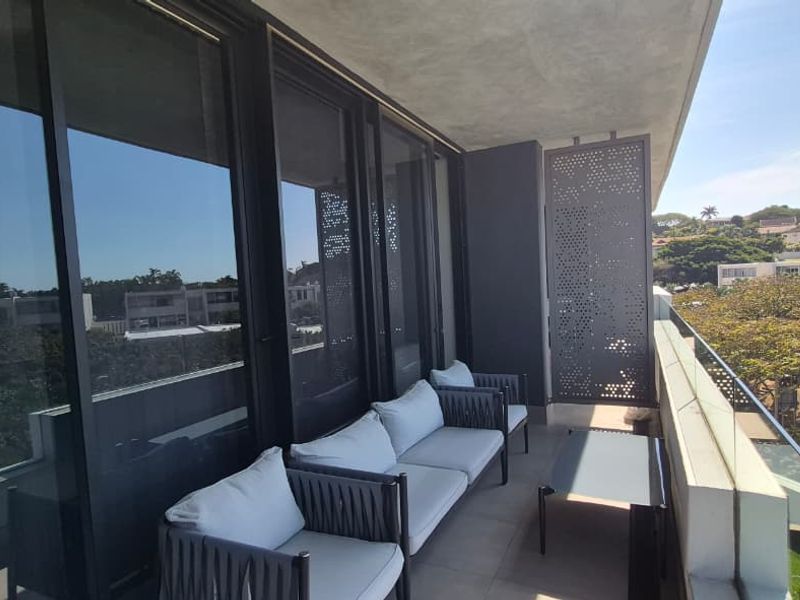 A modern covered balcony featuring a glass balustrade, sleek sliding doors, and a decorative perforated metal privacy screen overlooking the surrounding neighborhood.