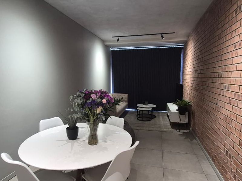Open plan living and dining area featuring a distinctive exposed brick feature wall, modern track lighting, and neutral tiled flooring.
