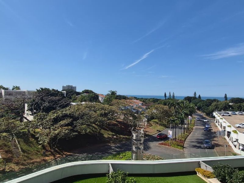 Elevated panoramic view overlooking the surrounding neighborhood, lush greenery, and distant ocean horizon from a balcony with a modern glass balustrade.