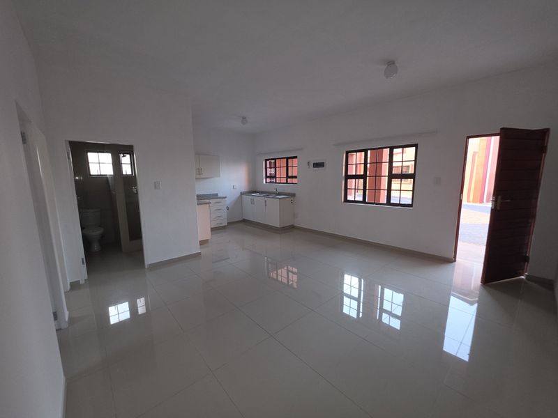 An open-plan living and kitchen area featuring tiled floors, neutral walls, and direct access to an adjacent bathroom and exterior entrance.