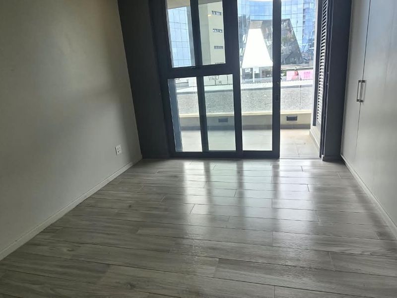 A bright, empty bedroom featuring modern laminate flooring, built-in cupboards, and a sliding glass door leading to a private balcony.