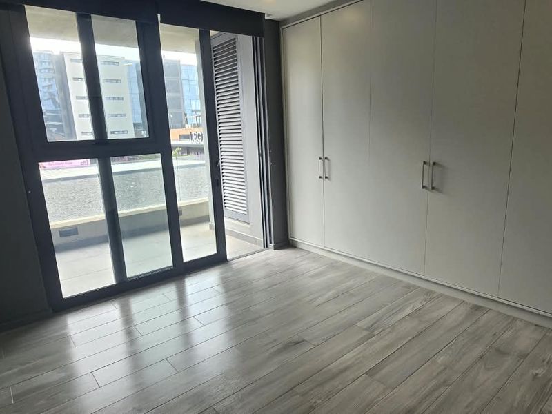 A bright, empty bedroom featuring modern tiled flooring, extensive built-in cupboards, and a large sliding glass door providing access to a private balcony.