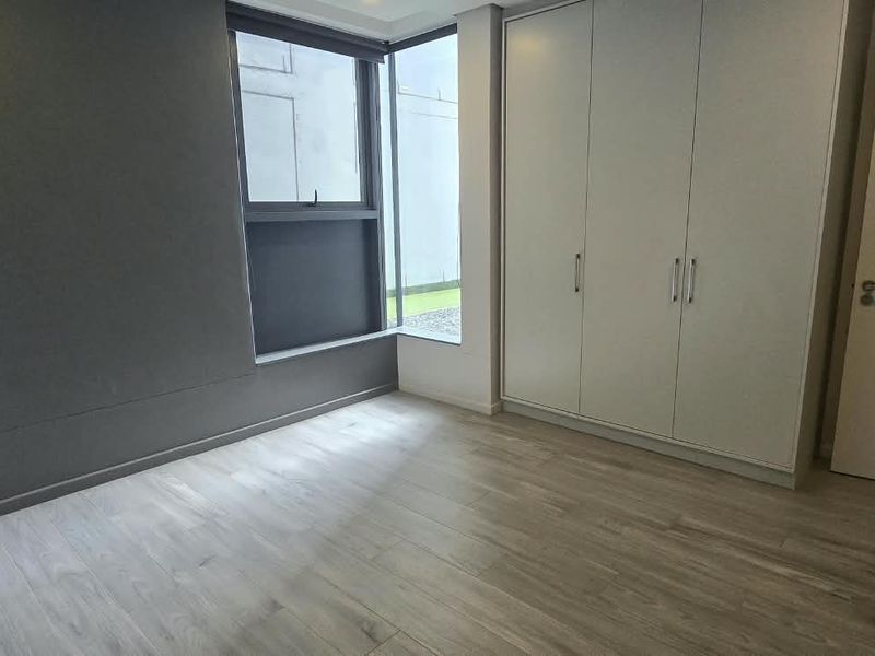 A bright, empty bedroom featuring modern laminate flooring, a large built-in wardrobe, and a corner window providing natural light.