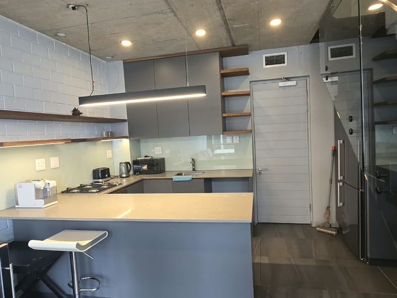 Modern kitchen featuring sleek grey cabinetry, stone countertops, gas hob, and floating timber shelves, set against an exposed concrete ceiling and brick wall.
