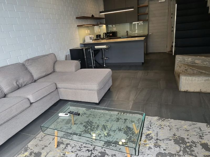 Modern open-plan living area featuring a contemporary kitchen with island, exposed brick walls, tiled flooring, and a staircase leading to the upper level.