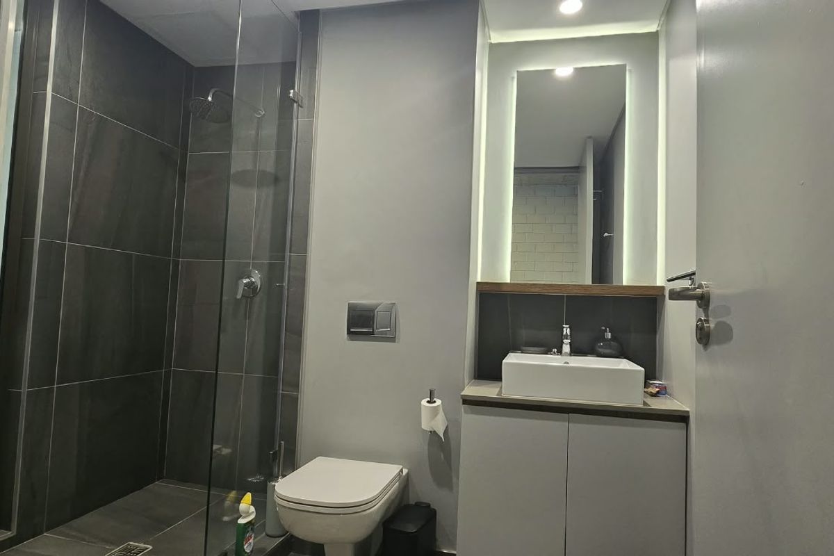 Modern bathroom featuring a walk-in shower with dark tiling, a wall-mounted toilet, and a vanity with a vessel sink and backlit mirror.