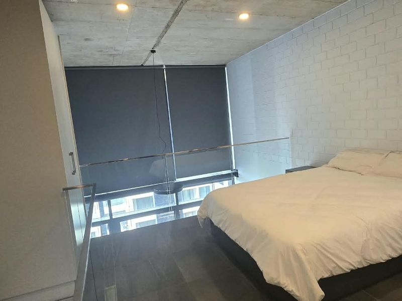 Modern loft-style bedroom featuring exposed concrete ceilings, painted brick walls, and a glass balustrade overlooking the lower level.