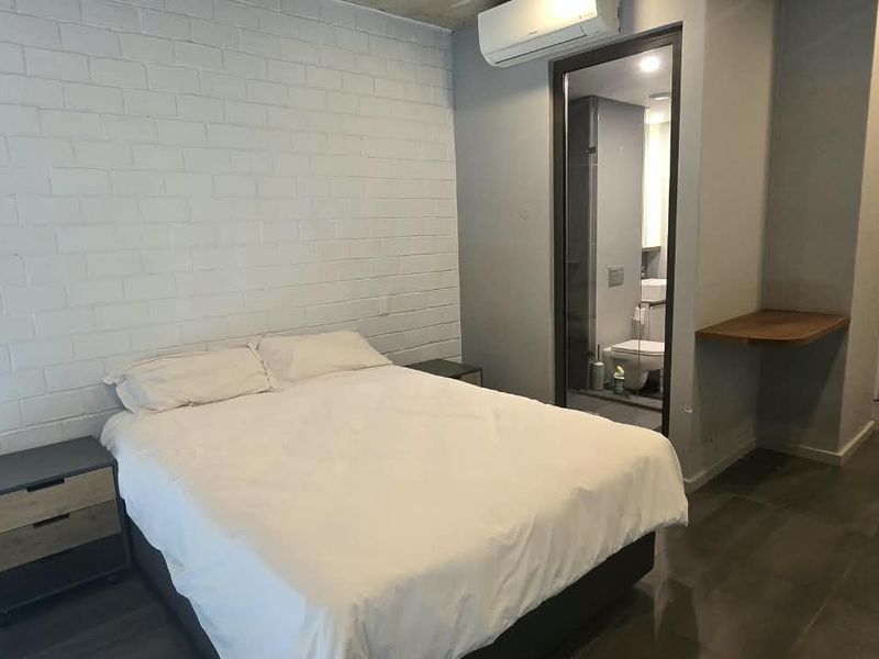 Modern bedroom featuring an exposed concrete ceiling, painted brick feature wall, split-system air conditioning, and an integrated en-suite bathroom.