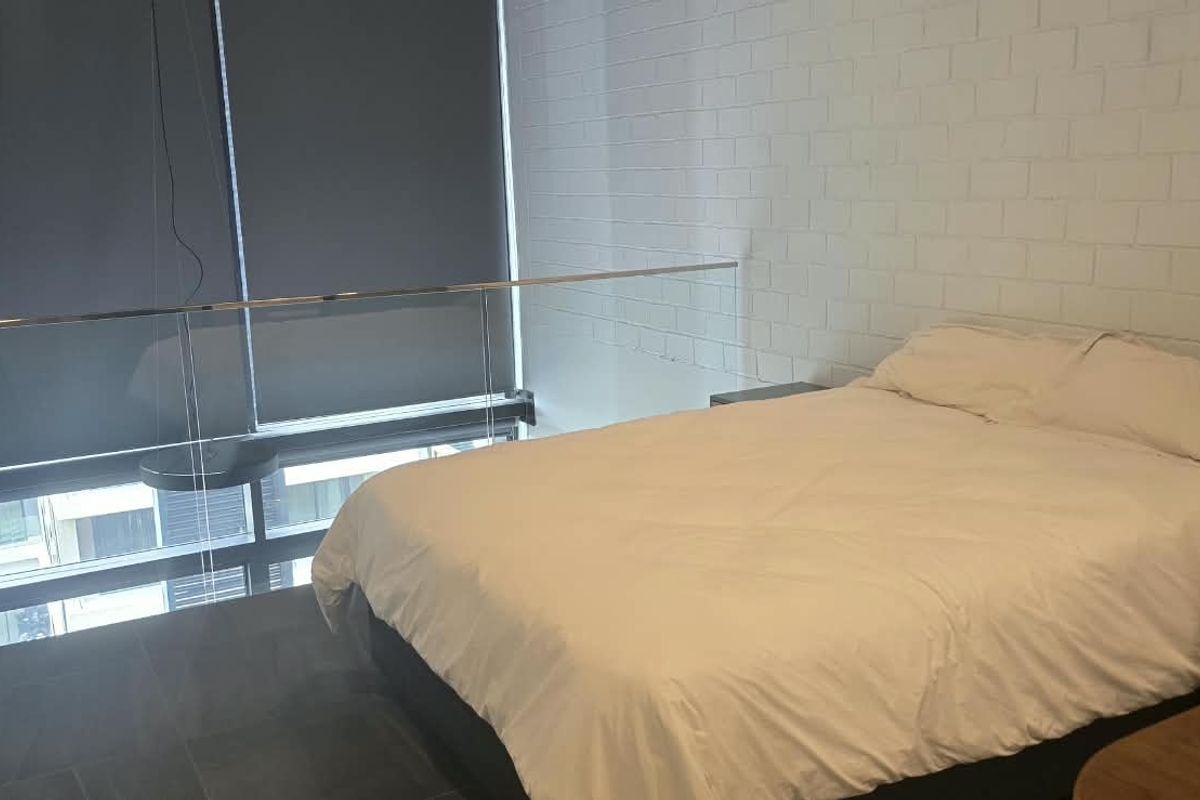 A mezzanine-style bedroom featuring an exposed brick wall, modern glass balustrade, and large windows with roller blinds.