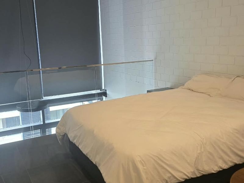 A mezzanine-style bedroom featuring an exposed brick wall, modern glass balustrade, and large windows with roller blinds.