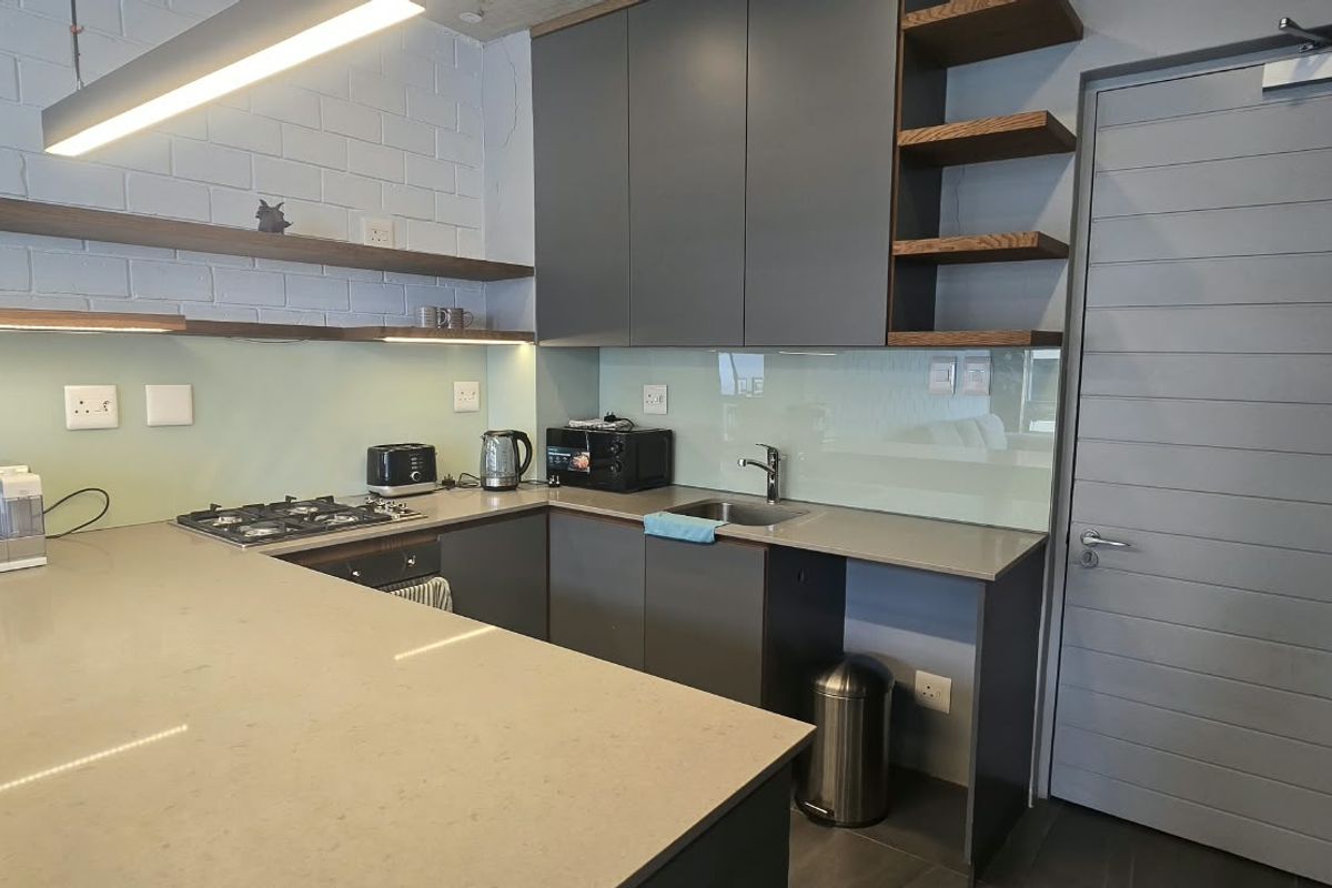 Modern kitchen featuring dark grey cabinetry, stone countertops, gas hob, under-counter oven, and stylish floating shelves with integrated lighting.